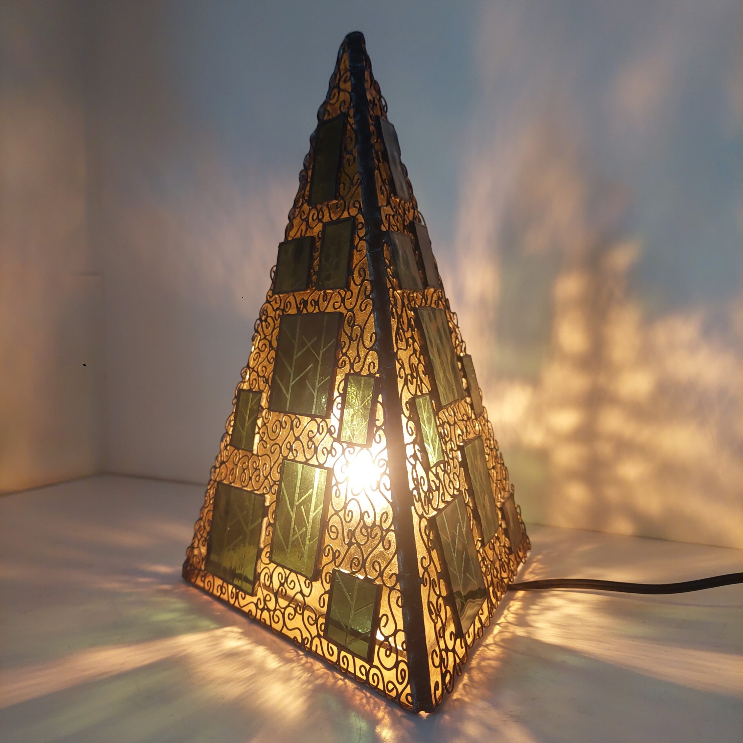 ステンドグラス、三角錐の置き型ランプです。秋の森をイメージし、黄色のガラスをベースに、緑のガラスにこの模様をサンドブラストしたガラスを重ねました。 細かくワイヤーを貼付け、蔓がランプをつたっているように見えます。