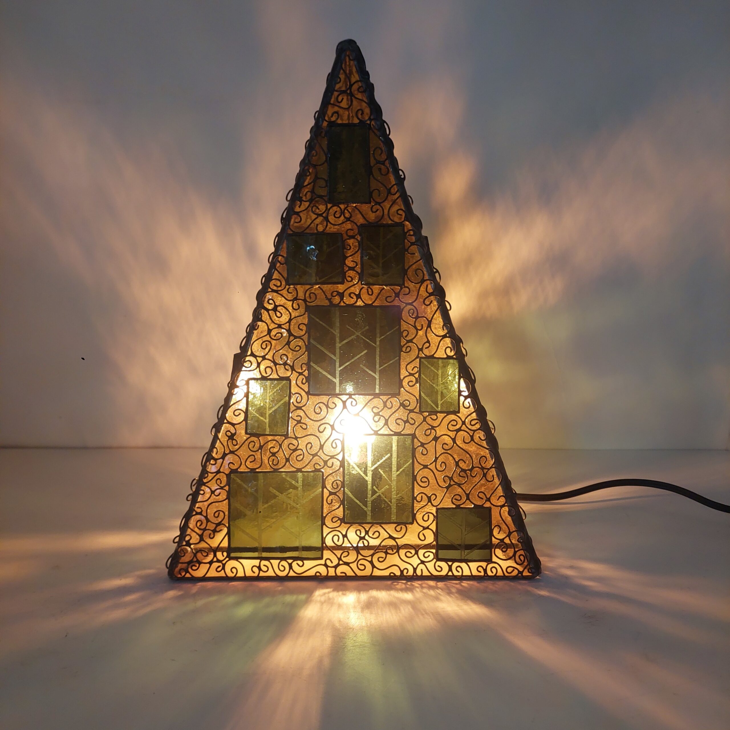 ステンドグラス、三角錐の置き型ランプです。秋の森をイメージし、黄色のガラスをベースに、緑のガラスにこの模様をサンドブラストしたガラスを重ねました。 細かくワイヤーを貼付け、蔓がランプをつたっているように見えます。