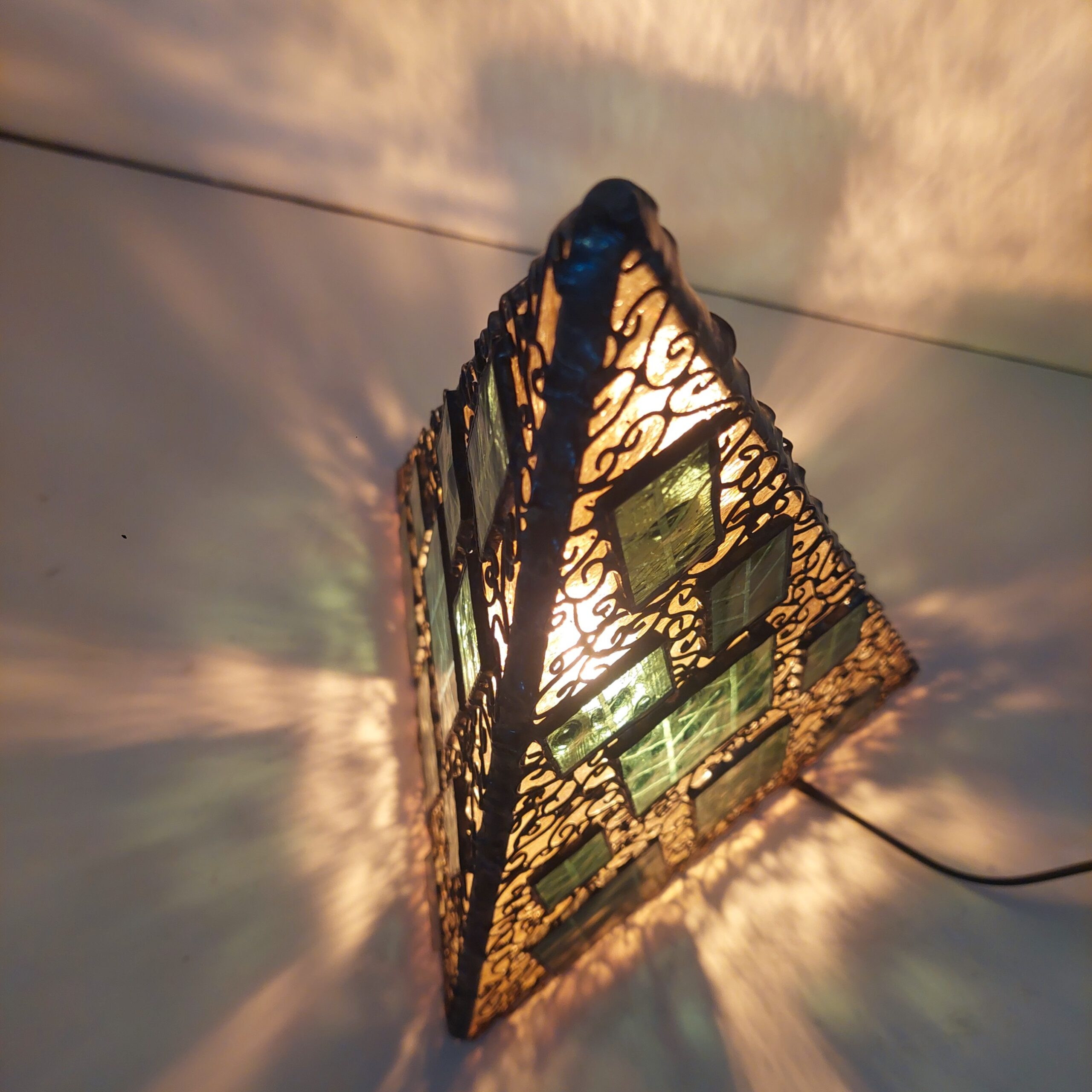 ステンドグラス、三角錐の置き型ランプです。秋の森をイメージし、黄色のガラスをベースに、緑のガラスにこの模様をサンドブラストしたガラスを重ねました。 細かくワイヤーを貼付け、蔓がランプをつたっているように見えます。