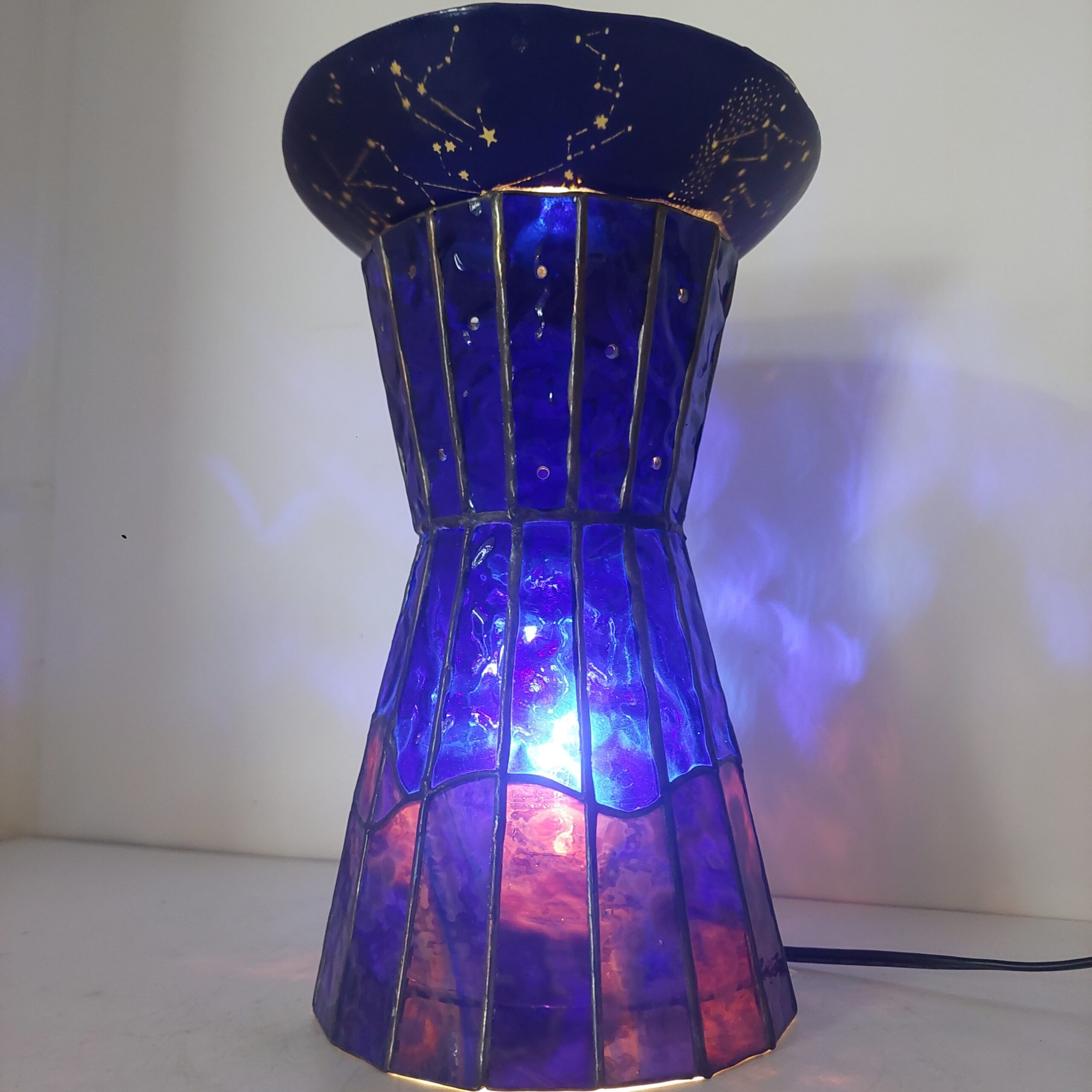 ステンドグラスの置き型ランプです。ランプ本体は赤~紫~青のガラスで夜の景色を表現、上には夜空をイメージし、沢山の星が彫られたお皿を乗せ、ライトアップします。まるで小さなプラネタリウムのような世界観のランプです。