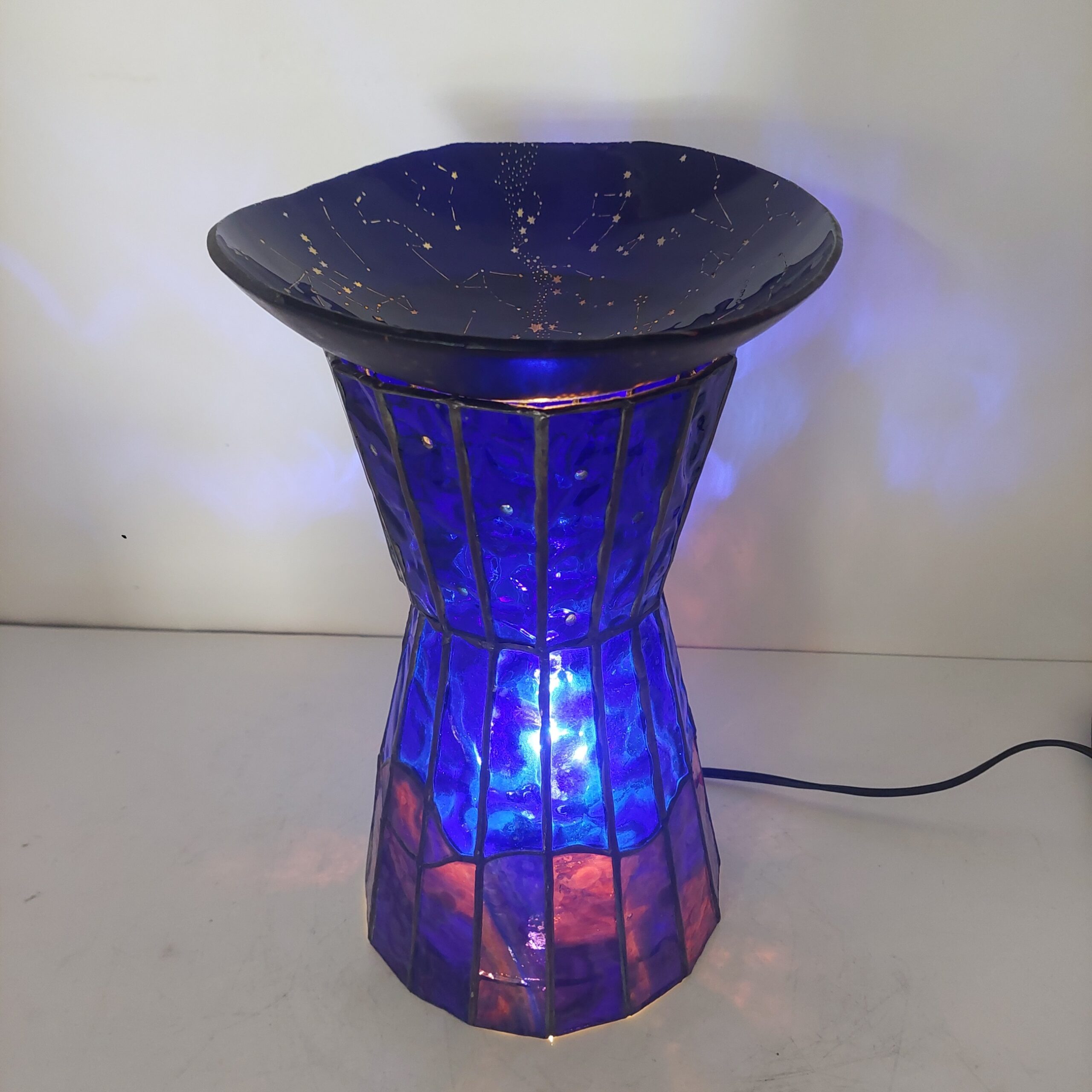 ステンドグラスの置き型ランプです。ランプ本体は赤~紫~青のガラスで夜の景色を表現、上には夜空をイメージし、沢山の星が彫られたお皿を乗せ、ライトアップします。まるで小さなプラネタリウムのような世界観のランプです。