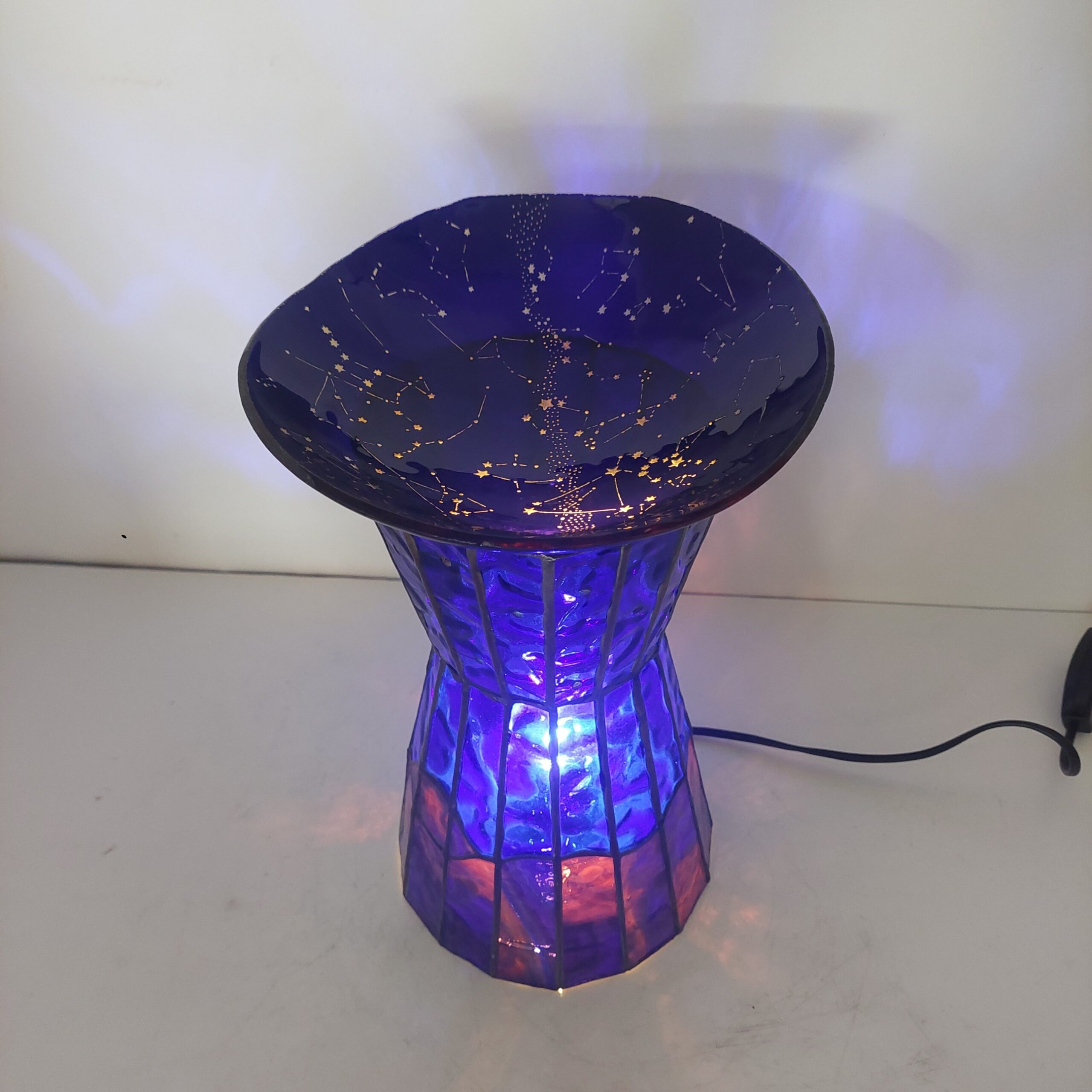 ステンドグラスの置き型ランプです。ランプ本体は赤~紫~青のガラスで夜の景色を表現、上には夜空をイメージし、沢山の星が彫られたお皿を乗せ、ライトアップします。まるで小さなプラネタリウムのような世界観のランプです。